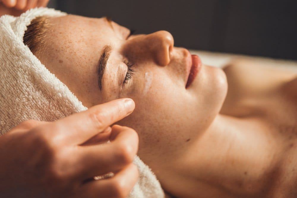 A person relaxes with their eyes closed as they receive a facial massage, wrapped in a towel - Skin Spa Health & Wellness A person relaxes with their eyes closed as they receive a facial massage, wrapped in a towel - Skin Spa Health & Wellness