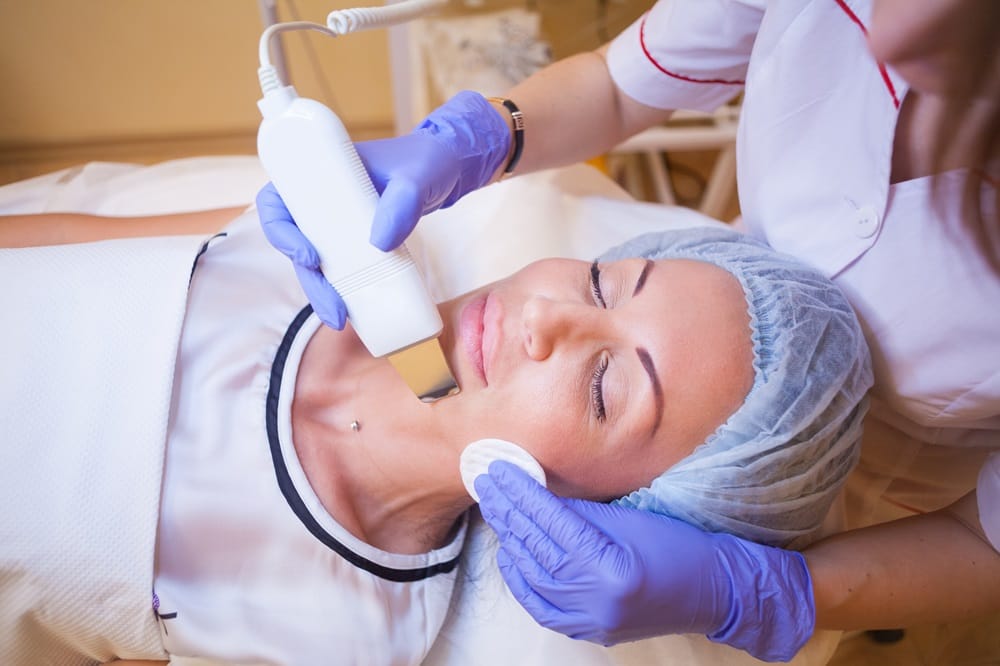 A woman receives a facial treatment with a handheld device, while a professional wearing purple gloves gently holds her face - Skin Spa Health & Wellness A woman receives a facial treatment with a handheld device, while a professional wearing purple gloves gently holds her face - Skin Spa Health & Wellness